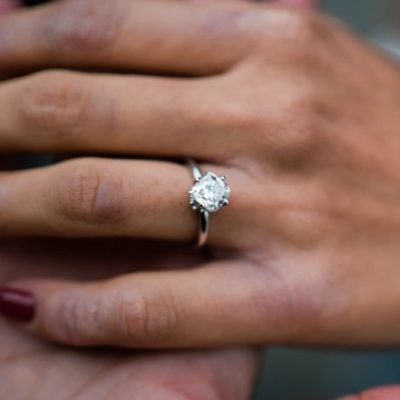 A woman's hand placed in a man's open one, featuring a cushion cut diamond offset to sit like a diamond with small diamonds sitting below basket to appear like a bouquet, and a white gold band.