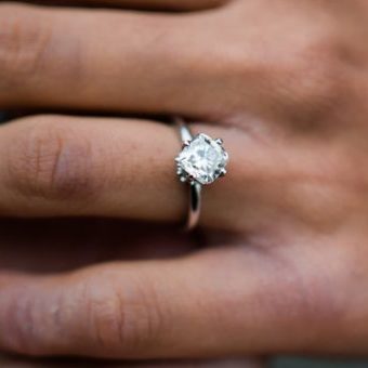 A woman's hand placed in a man's open one, featuring a cushion cut diamond offset to sit like a diamond with small diamonds sitting below basket to appear like a bouquet, and a white gold band.