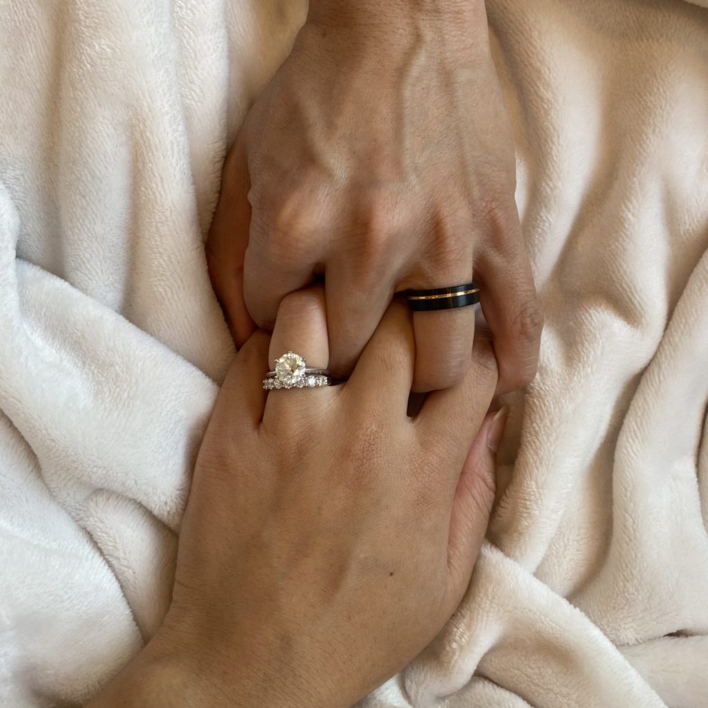 A couple's interlocked hands, his with a black and gold tungsten band and hers with a diamond engagement ring and matching diamond wedding band.