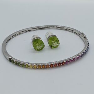 A white gold bangle style bracelet with half covered in a rainbow of sapphires ranging from green through to blue, and large oval peridot studs that matches a green portion of the sapphire rainbow.