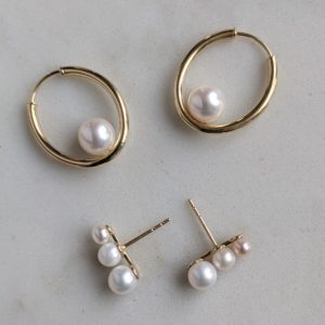 Oval hoops with large pearls affixed to the inner portion of the base of the oval, and graduated pearl climbers studs with 3 pearls each, both set in yellow gold, on a white marble background.