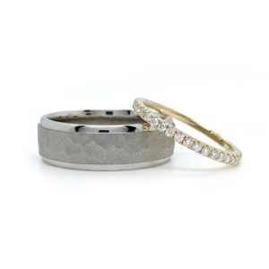 Men's white gold wedding band with a thick sandblasted hammered finished section that is set between two thinner polished ends. A women's thin yellow gold pave diamond band rests on it.