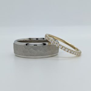 Men's white gold wedding band with a thick sandblasted hammered finished section that is set between two thinner polished ends. A women's thin yellow gold pave diamond band rests on it.