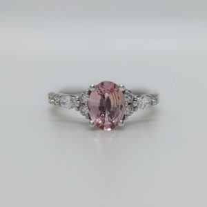 Oval padparasha and diamond engagement ring with a round cluster and marquise diamonds on either side of the centre stone and a pave band set in white gold.