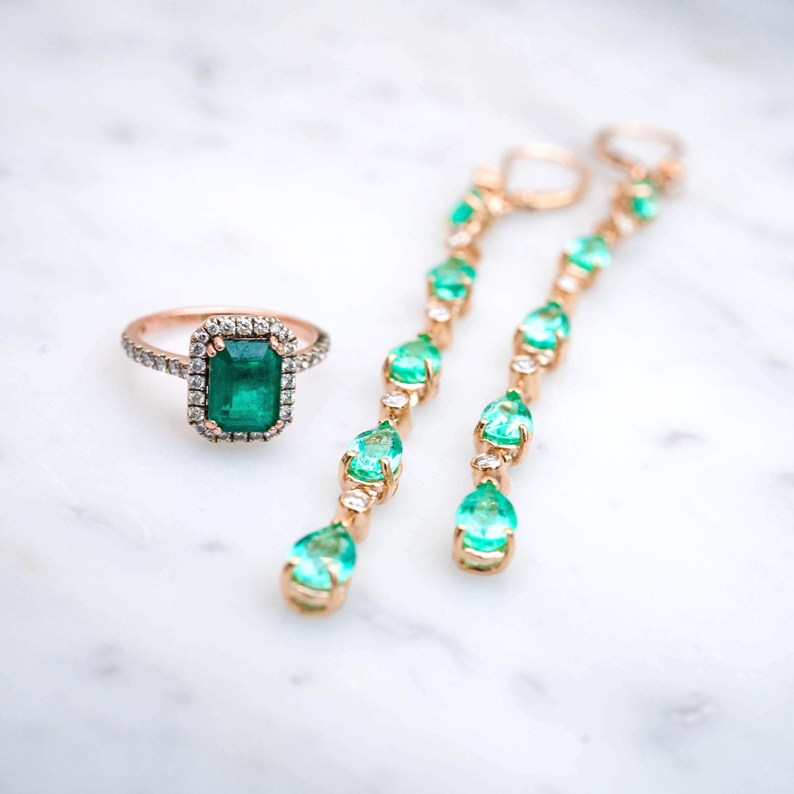 Emerald engagement ring set in white and rose gold beside a pair of 6 pear shaped emerald & diamond drop earrings also set in rose gold.